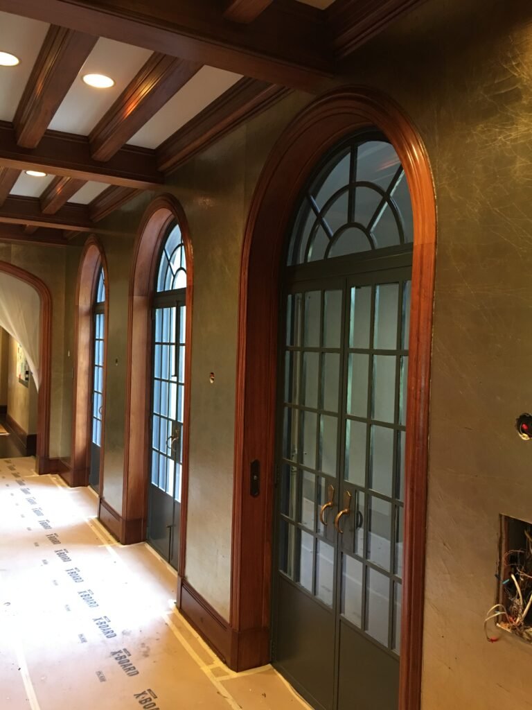 Hallway with textured metallic-finish plaster walls and arched wood-trimmed windows and doors, floor covered in protective paper during a wallcovering installation