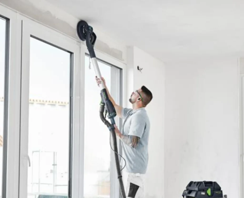 Worker using a long-reach sander with a dust extraction vacuum to smooth a drywall seam near the ceiling.