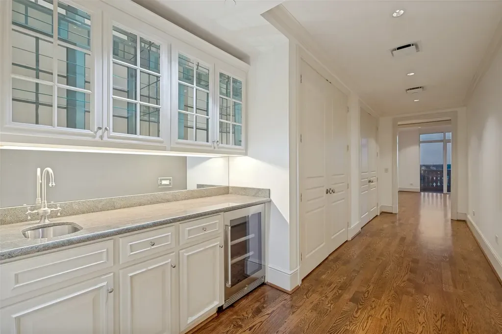 White painted wet bar cabinets with glass-front uppers, granite countertop, and an adjacent hardwood floor hallway.