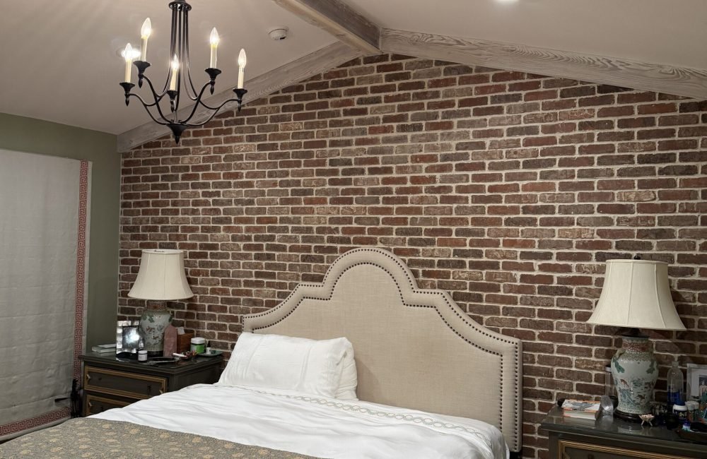Bedroom with a realistic brick-pattern wallpaper accent wall behind an upholstered headboard, wrought iron chandelier, and matching nightstand lamps