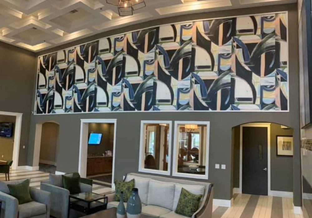 Two-story lobby with a bold abstract geometric wallpaper mural above dark gray walls, coffered ceiling with chandelier, and a seating area with gray sofas below
