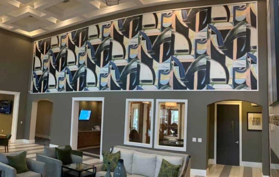 Two-story lobby with a bold abstract geometric wallpaper mural above dark gray walls, coffered ceiling with chandelier, and a seating area with gray sofas below