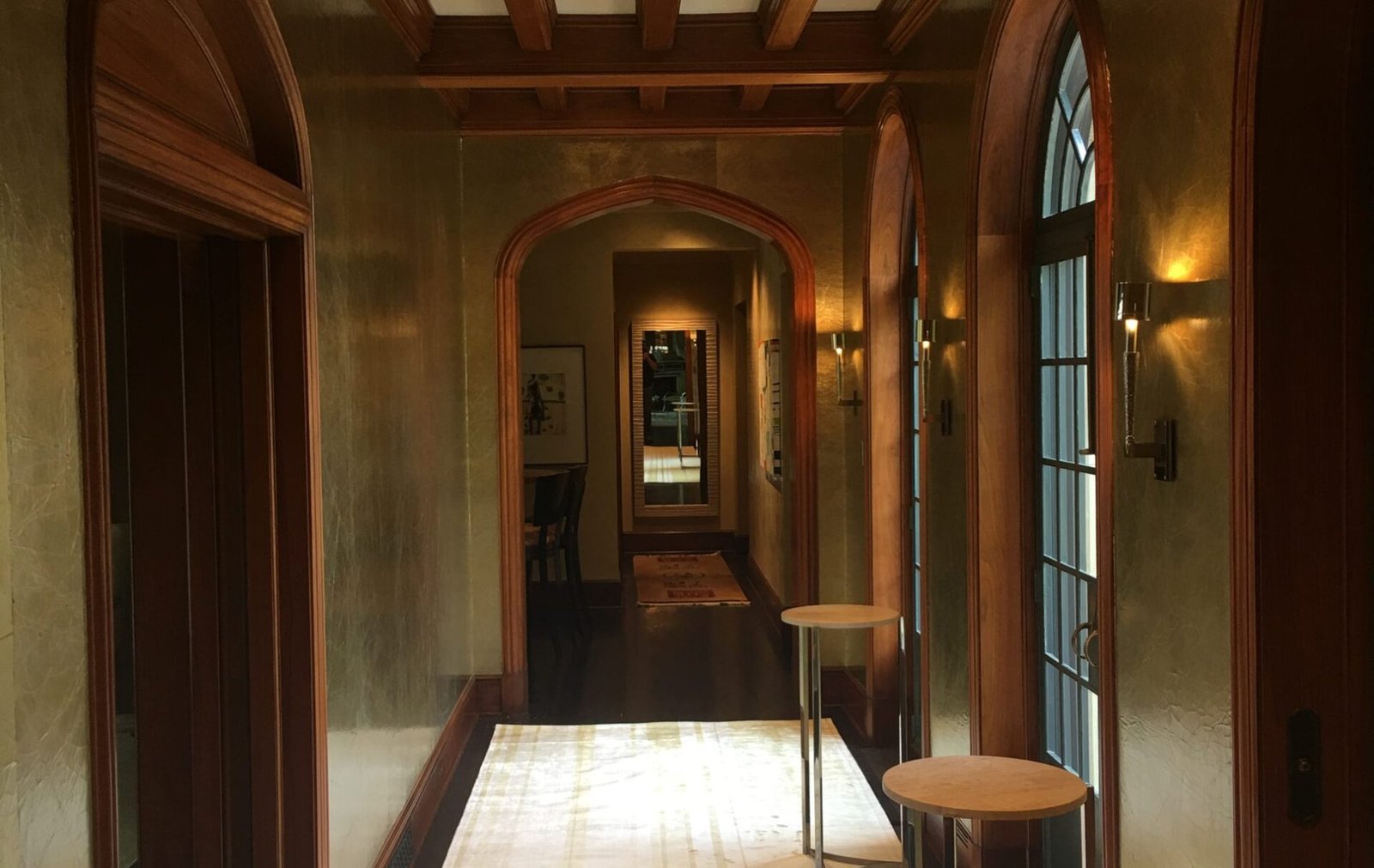 Arched hallway with textured plaster walls, dark wood trim and beamed ceiling, leading to a foyer with a runner rug and mirrored console table