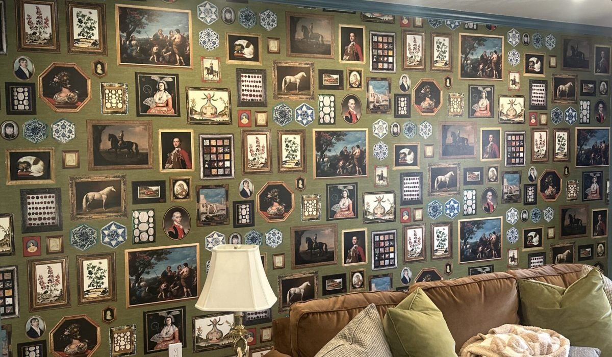 Olive-green feature wall wallpaper printed with a gallery-style pattern of ornate framed portraits, horses, and decorative medallions, above a brown leather sofa with green and cream pillows