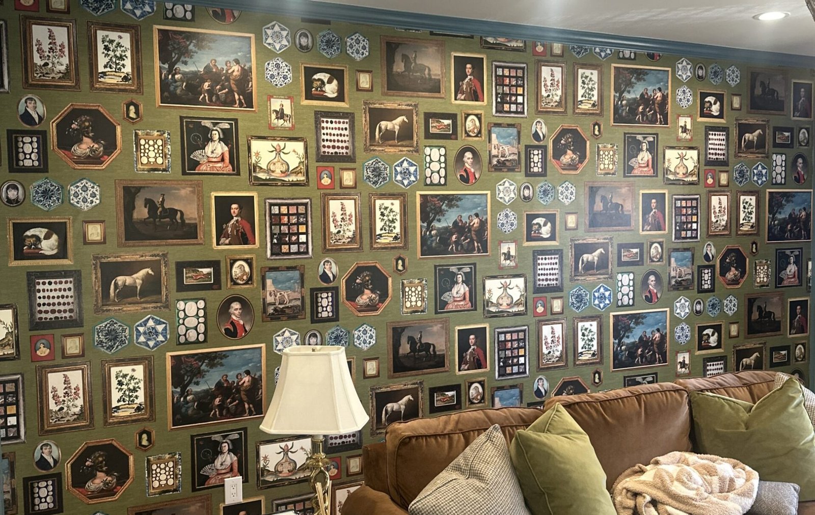 Olive-green feature wall wallpaper printed with a gallery-style pattern of ornate framed portraits, horses, and decorative medallions, above a brown leather sofa with green and cream pillows